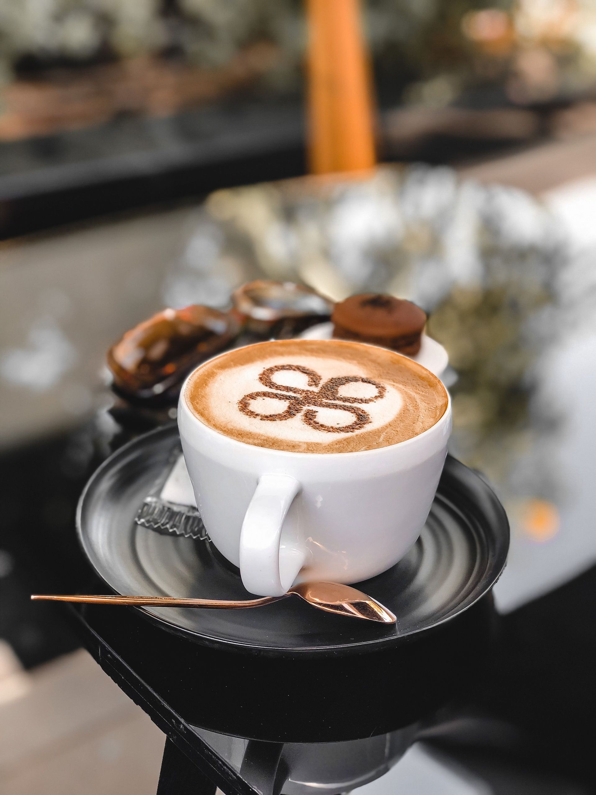 Close-up of a cappuccino with latte art on a black saucer with a teaspoon, shot outdoors.