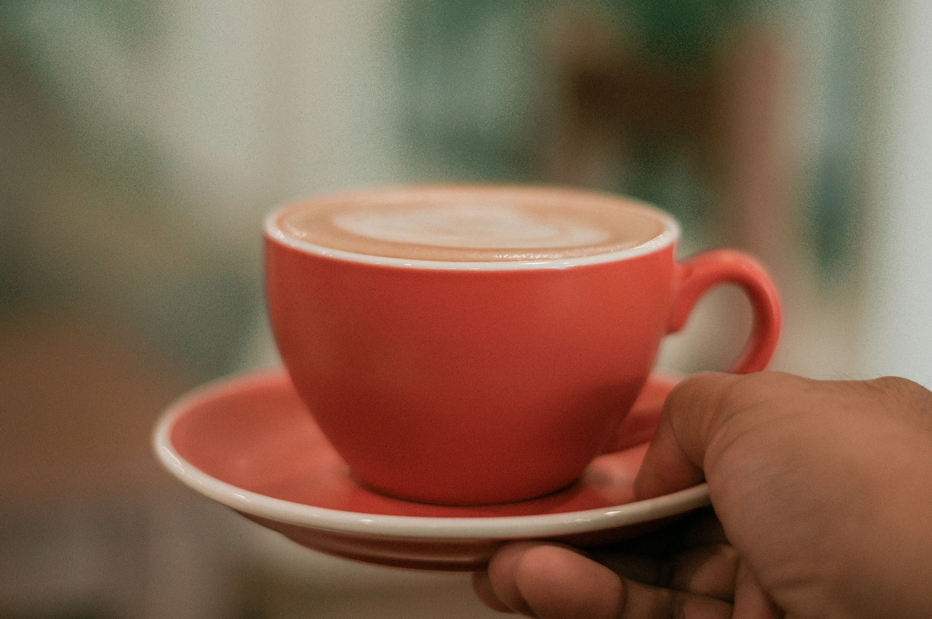 Crop unrecognizable person with red mug of hot coffee with latte art in hand on blurred background in light cafe