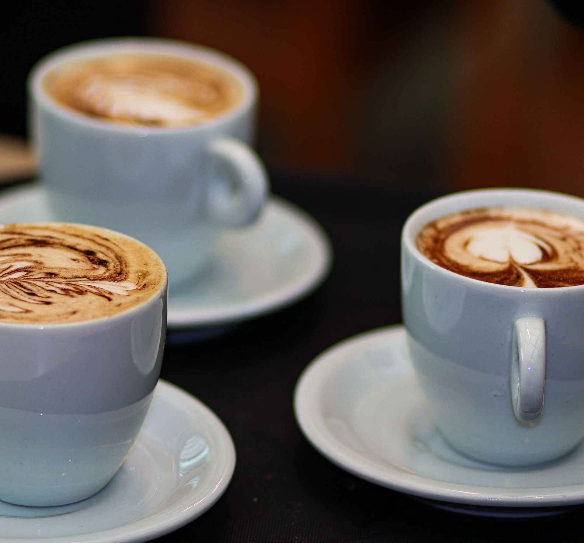 Three cappuccinos with intricate latte art in white cups on saucers, showcasing coffee artistry.
